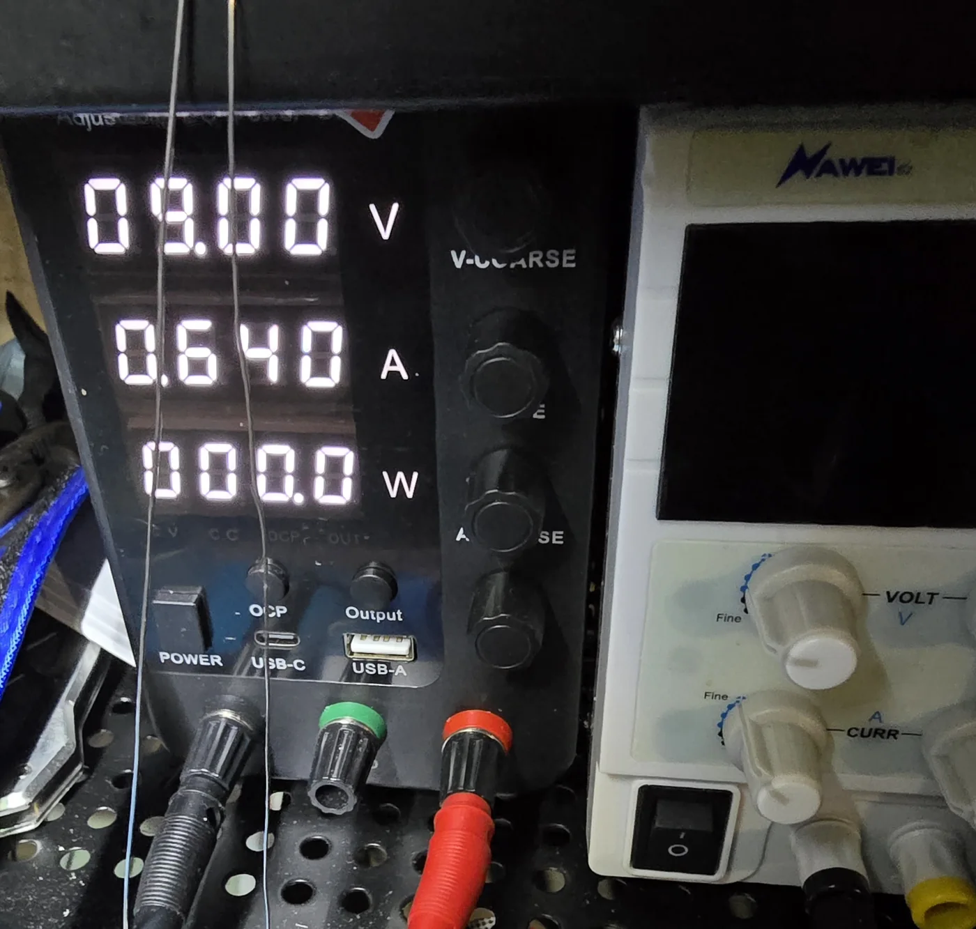 Two bench power supplies side by side, one black showing 9.00 V and 0.640 A on its display with three output terminals, one cream-coloured with fine voltage and current knobs and a front rocker switch.