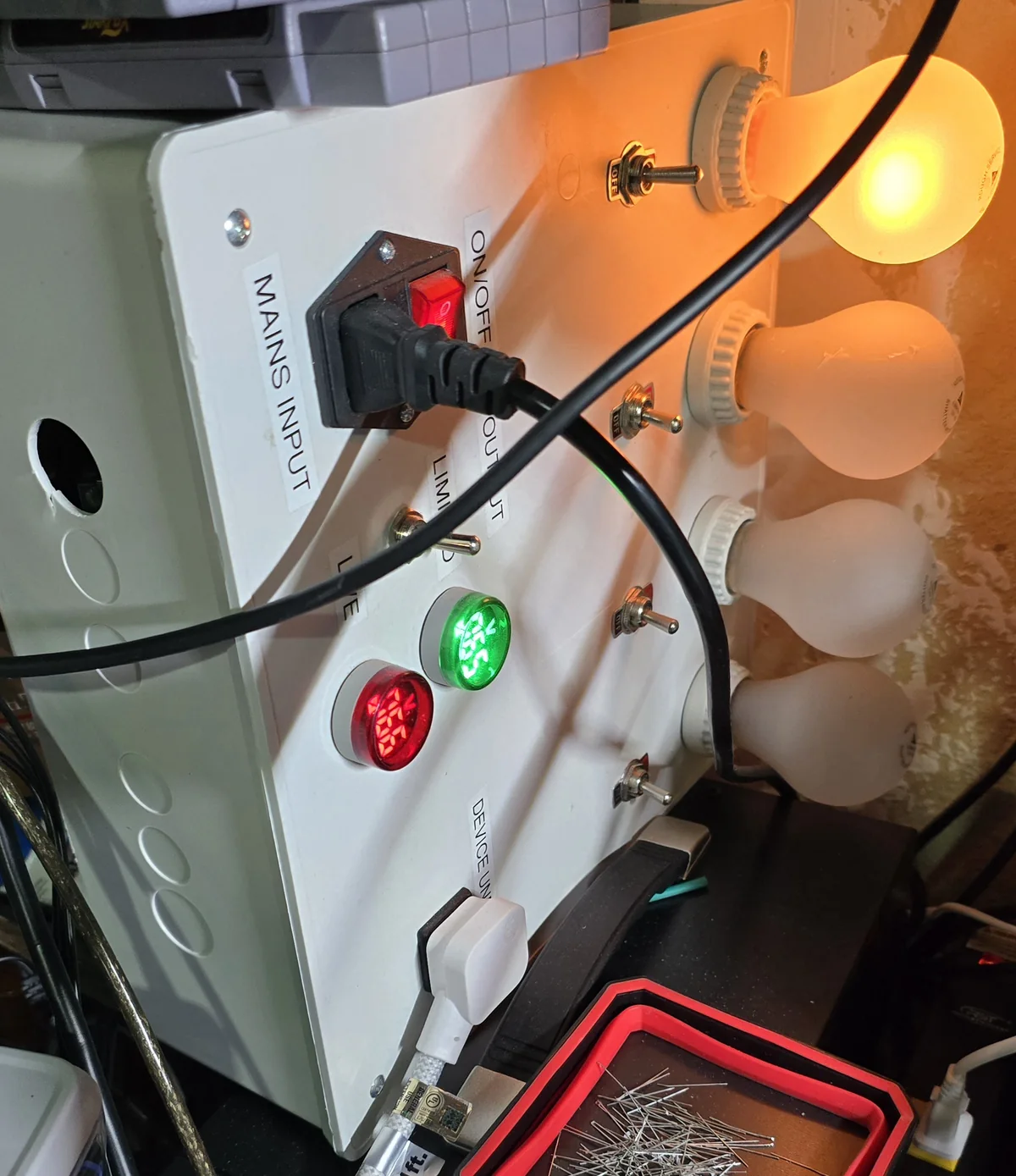 A home-built dim bulb tester in a white enclosure with four bulb sockets, toggle switches and AC meters, with one bulb lit bright orange.