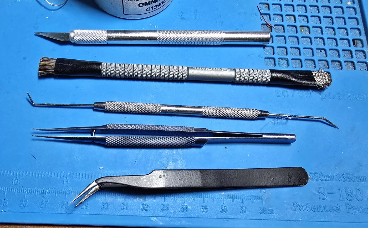 A dental pick, a stiff brush for cleaning flux, an X-Acto knife and two pairs of tweezers laid out on a work mat.