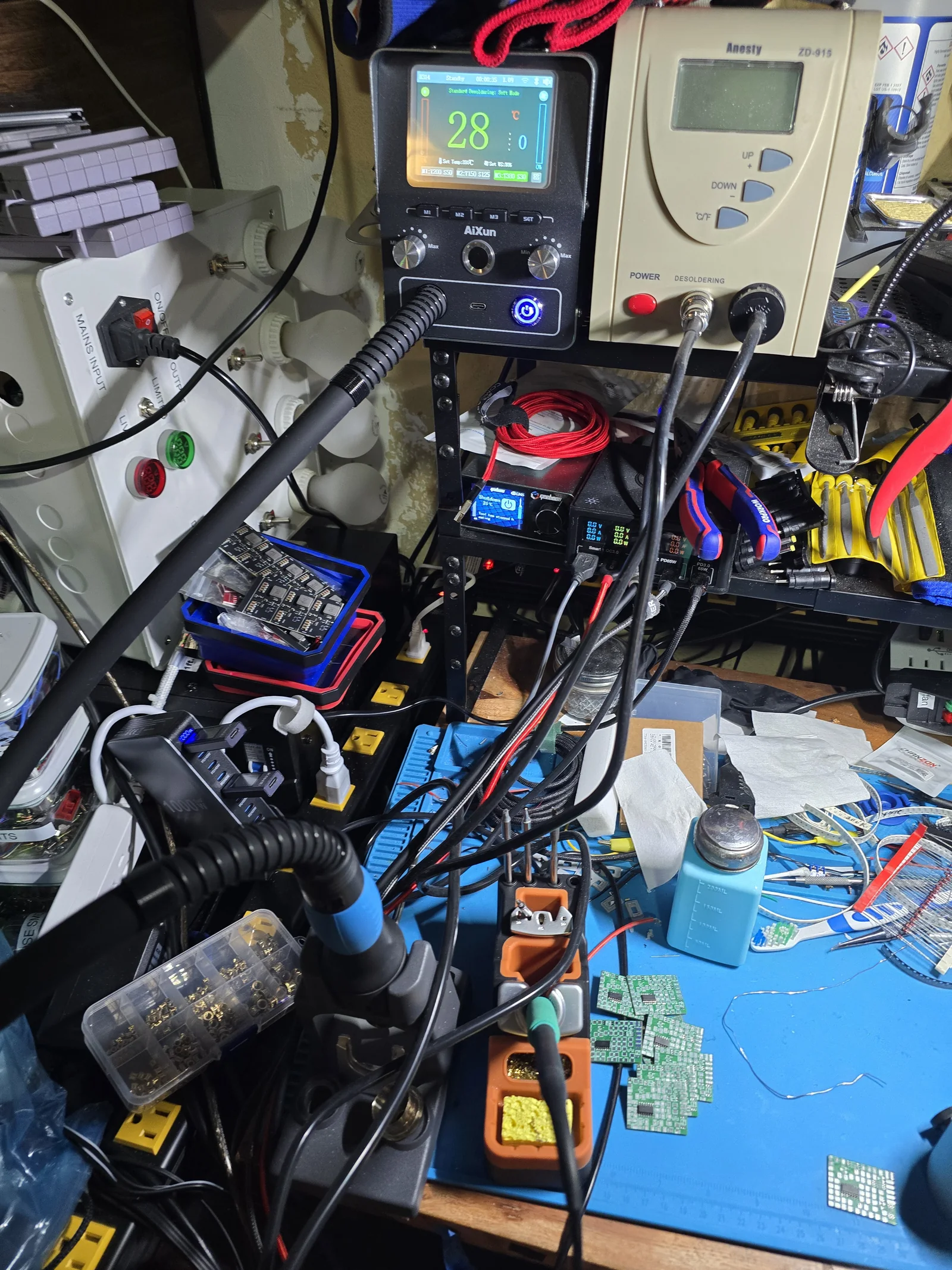 A cluttered electronics workbench with an AiXun hot air station in the foreground, a desoldering station above it, and a dim bulb tester below.