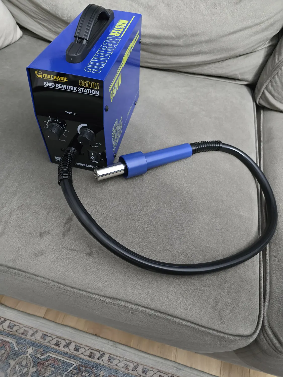 A blue Mechanic 957DW SMD rework station standing upright on a grey sofa cushion, with a black carry handle across the top. The black front panel is printed SMD REWORK STATION and 957DW and carries two black knobs, one labelled TEMP in degrees Celsius with a scale numbered 2 to 8 and one labelled 450 degrees C Control, plus a rocker switch marked ON and OFF beside the word POWER. A thick black corrugated air hose leaves the front of the base and curves across the cushion to a blue handpiece with a chrome nozzle.