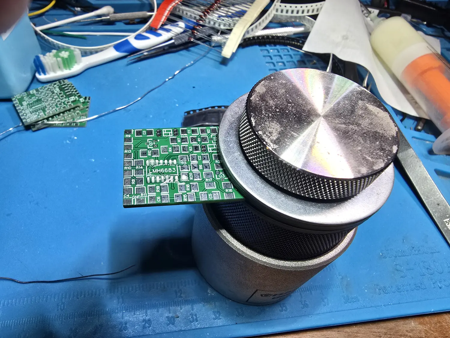 A Hakko Omnivise on a blue silicone bench mat printed with a centimetre ruler. The holder is a heavy machined metal cylinder with a knurled band around it and a flat disc clamp on top, gripping the edge of a small green Torapu SNES RGB mod board silkscreened LMH6683 with numbered resistor and capacitor pads. Behind it on the mat are tweezers, taped strips of axial resistors, a toothbrush, a flux bottle and two more small green boards.