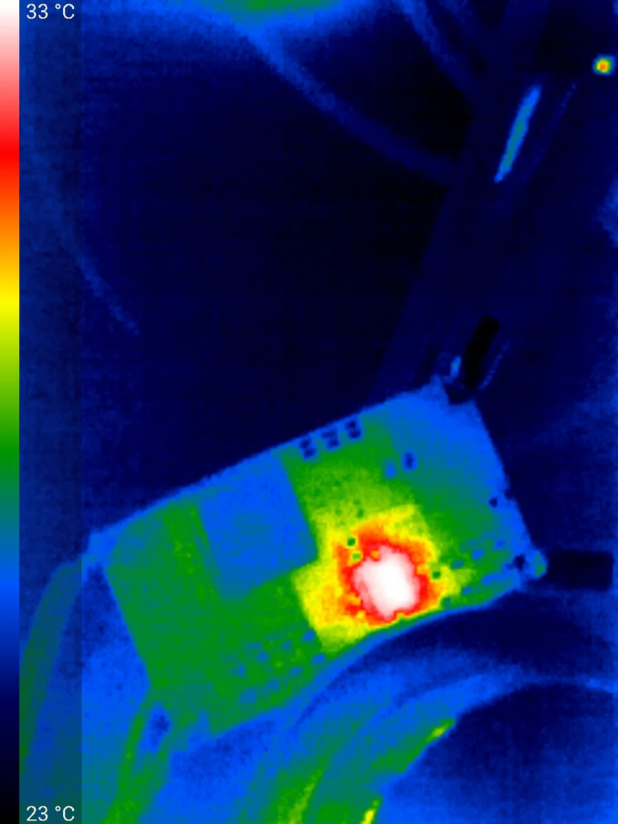 Thermal camera image of a circuit board on a 23 to 33 degree Celsius scale, with one component glowing white and red against a cool green and blue board.