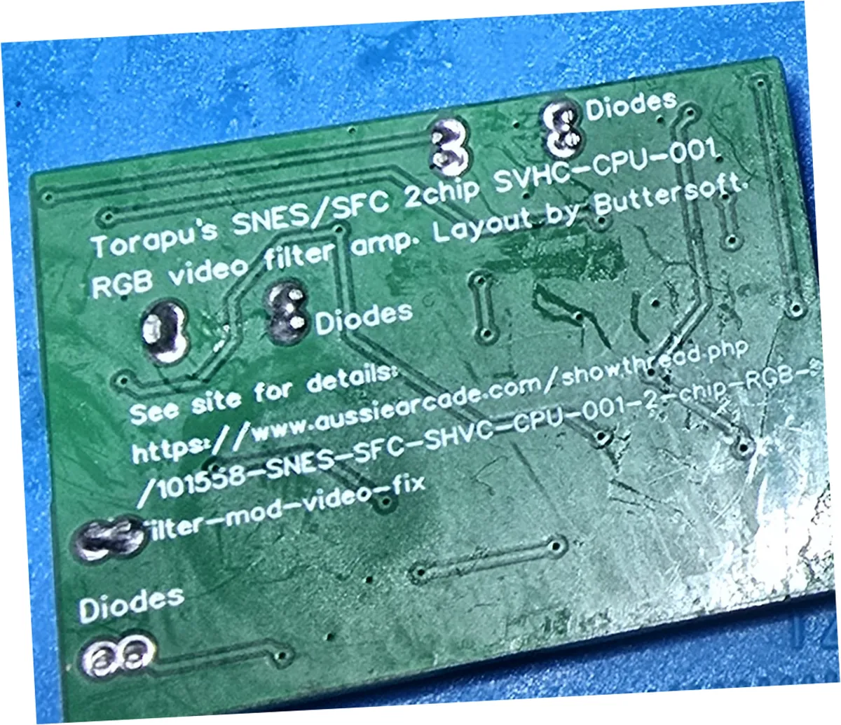 Close-up of the smooth solder side of a green circuit board, silkscreened Torapu's SNES slash SFC 2chip SVHC-CPU-001, RGB video filter amp, Layout by Buttersoft, with an aussiearcade forum URL and several pads labelled Diodes. A milky white film of dried flux is smeared across the solder mask, heaviest along the right-hand edge and streaked between the traces.