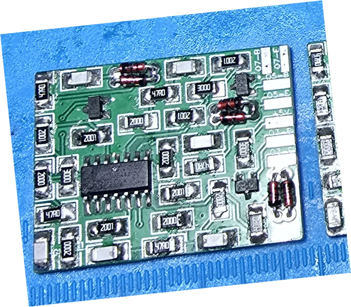 Close-up of the component side of the assembled RGB video filter amp board on a blue bench mat before cleaning. Dozens of surface-mount resistors marked 2001, 2000, 3000 and 47R0 surround a black 14-pin SOIC op-amp, with three pairs of axial diodes along the right-hand edge. The solder joints are dull and grey and a whitish crust of dried flux residue sits around the diode leads and across the pads.
