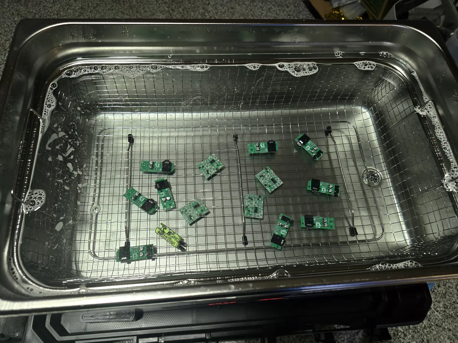 Looking down into the stainless steel tank of a 30 litre ultrasonic cleaner, filled with cleaning solution and fitted with a wire mesh basket. About a dozen small green circuit boards are spread across the basket, several with black connectors fitted. Foam has gathered along the tank rim and a drain fitting is visible at the right-hand end.