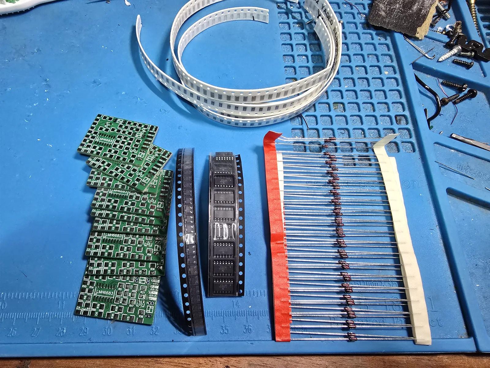 Bare green PCBs fanned out on a blue bench mat next to cut tape of SOIC-14 op-amps, a strip of 1206 passives, and a bandolier of axial 1N4148 diodes.