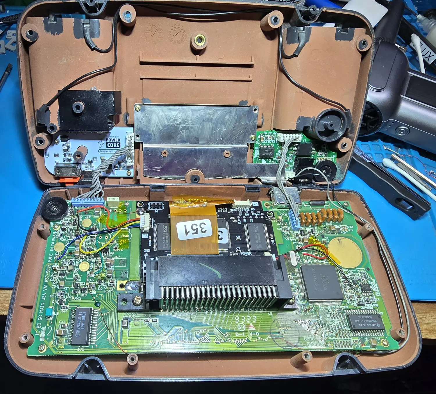 The Game Gear opened on the bench, both shell halves apart, showing the IPS driver board over the cartridge slot and the RetroSix PowerCore board in the rear shell