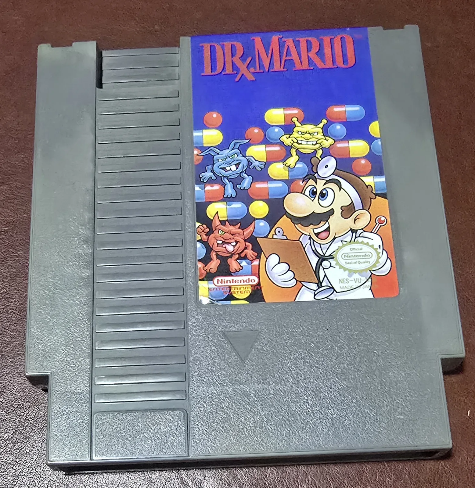 Front of an authentic Nintendo Dr. Mario cartridge for the NES, Dr. Mario tossing megavitamins at the red, blue, and yellow viruses with the official Nintendo Seal of Quality