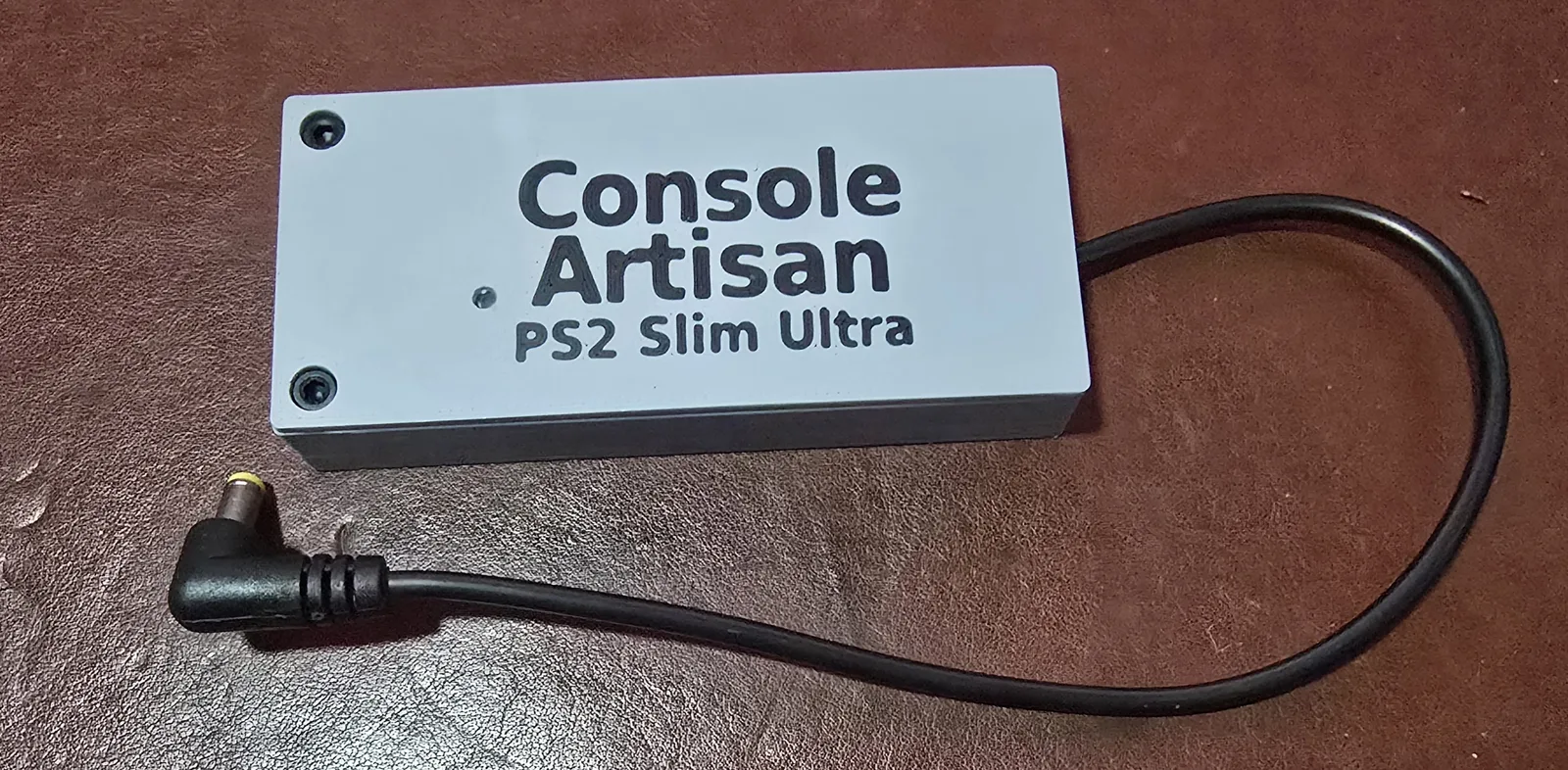 The finished Console Artisan PS2 Slim Ultra power supply: a compact white and grey printed case with the model name on the lid, its black output cable coiled beside it ending in a right-angle barrel plug