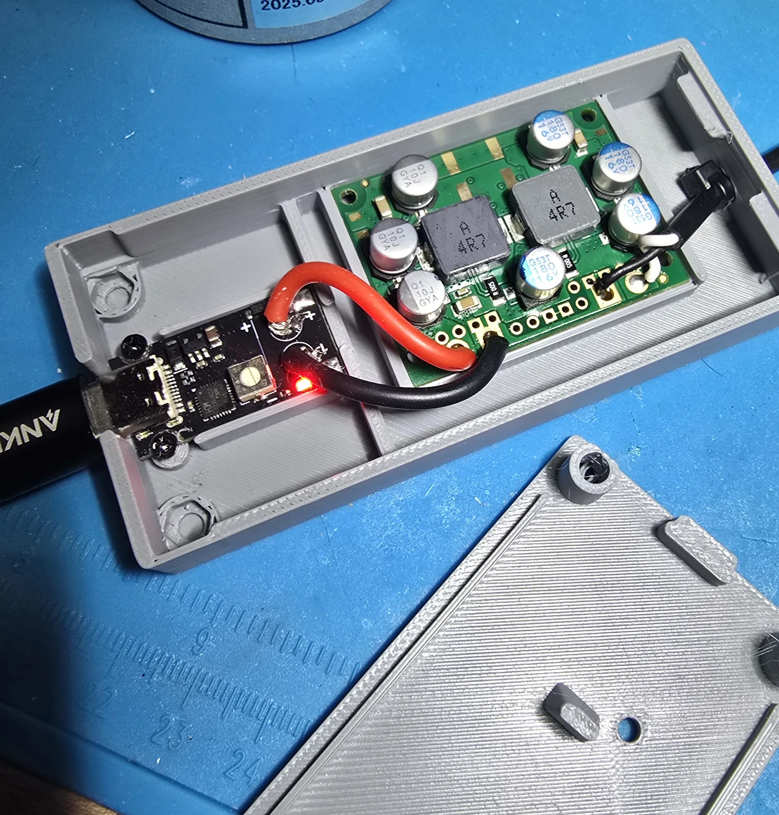 The case open and powered, showing the USB-C trigger board wired to the buck converter with heavy red and black wire, the trigger's LED lit, and the lid resting beside it
