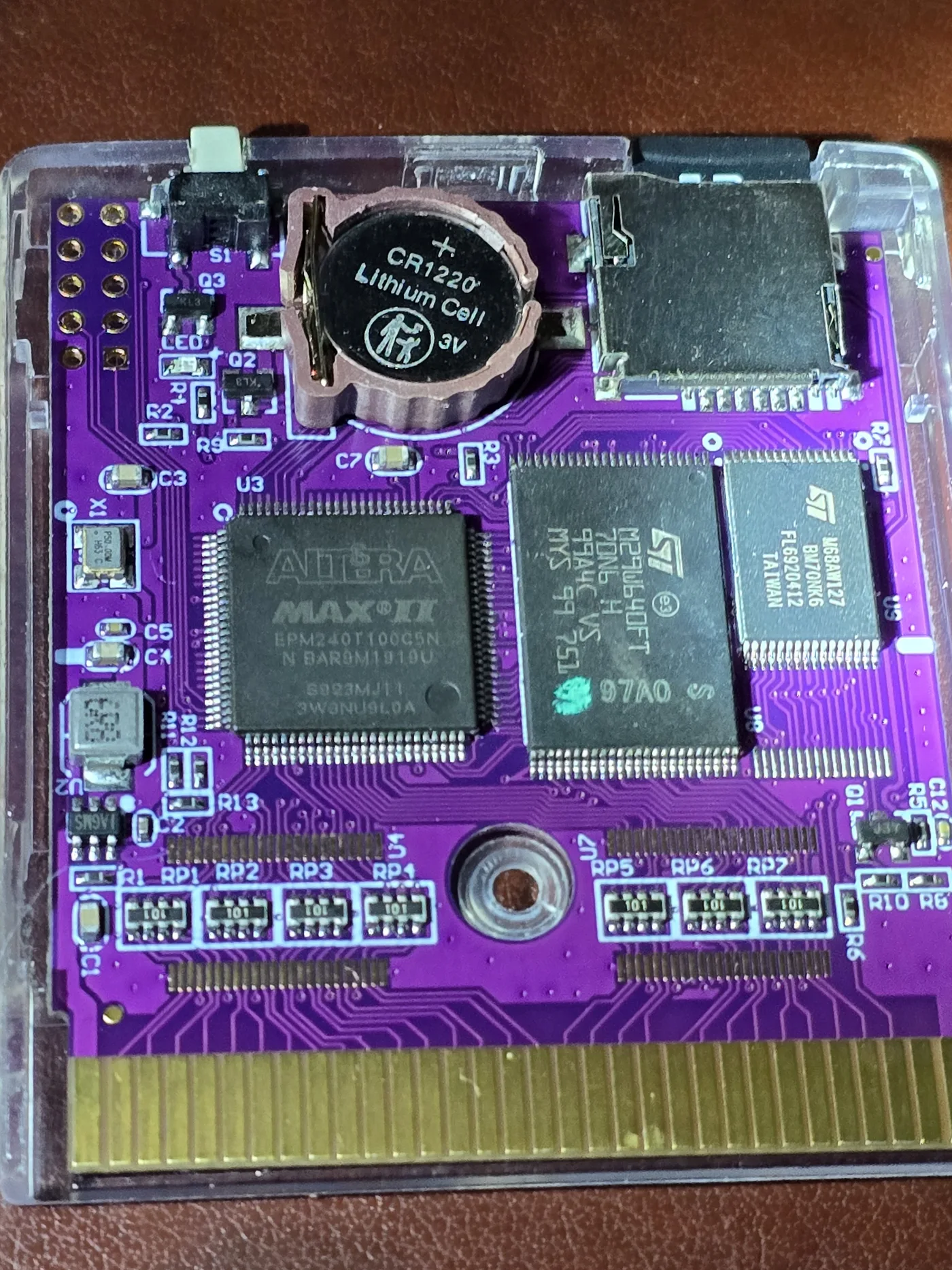 Component side of the GB Pro+ flash cartridge PCB lit from a low angle, showing an Altera MAX II EPM240T100C5N at U3, an ST M29W640FT at U8 and an ST M68AW127 at U9, a CR1220 lithium cell in a clip, a microSD socket, a tactile switch at S1, seven resistor networks marked 101 and an empty two-by-five plated header footprint.