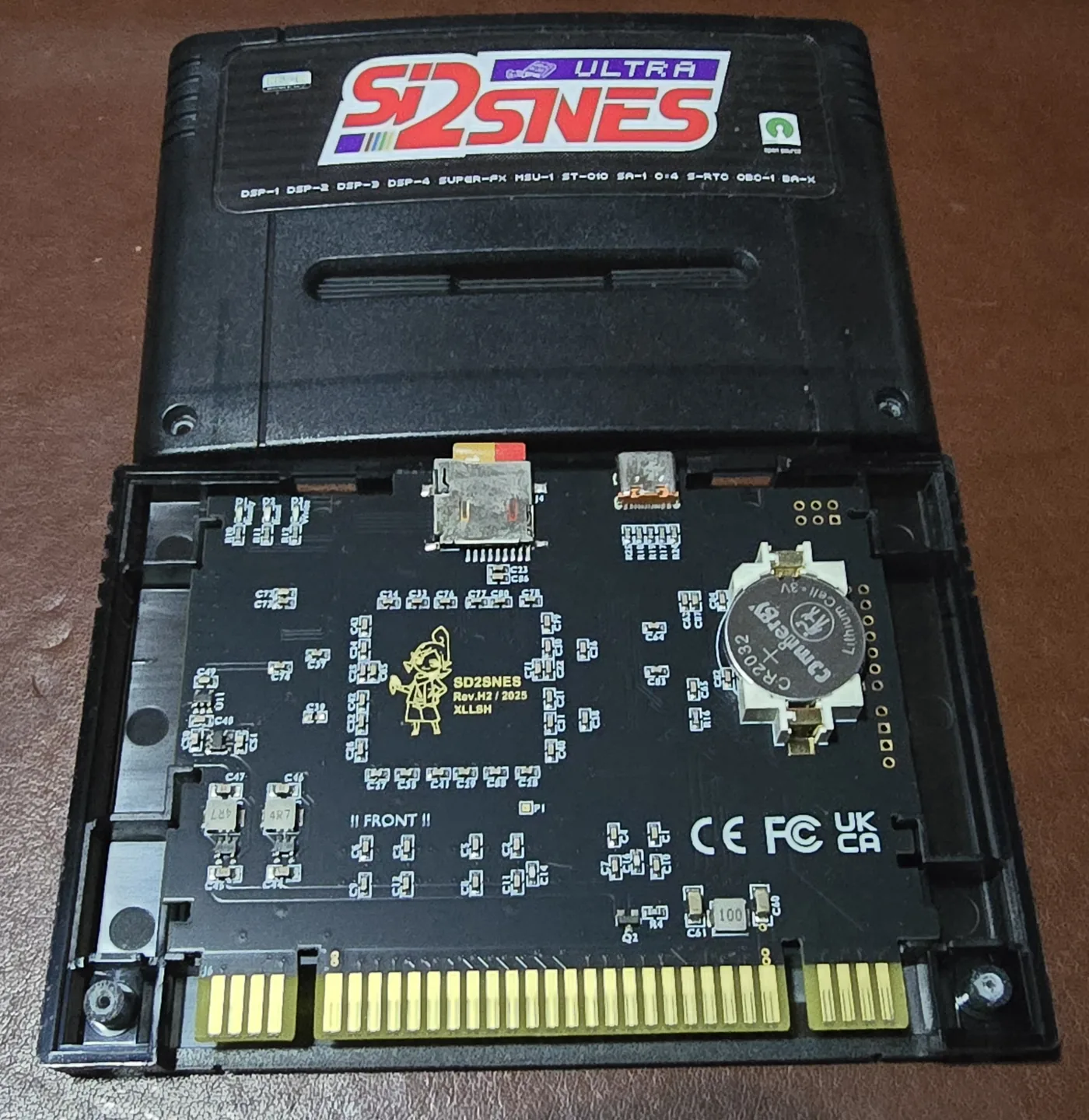 SD2SNES Ultra cartridge in a black shell with the back cover removed, showing a densely populated PCB with a CR2032 coin cell, microSD slot and USB-C port.