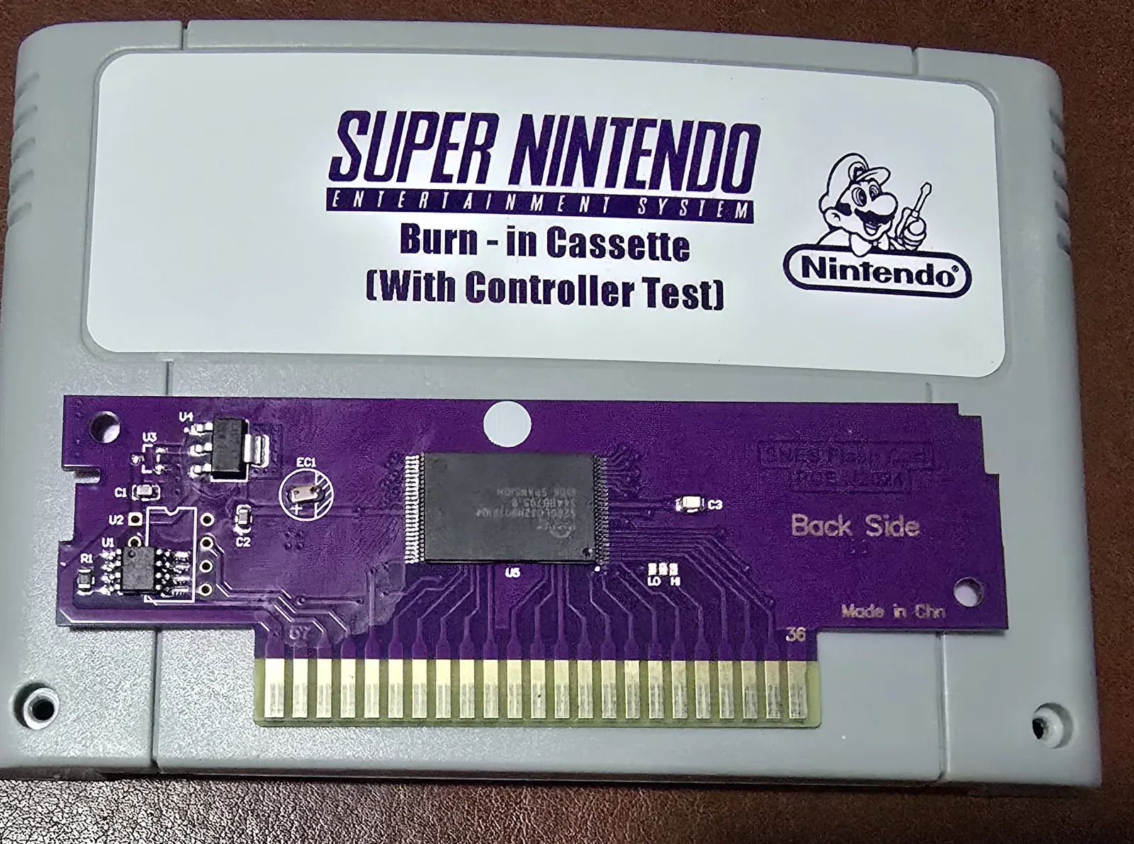 A grey Super Nintendo cartridge with a counterfeit Nintendo-branded label reading Burn-in Cassette (With Controller Test), shell opened to expose a purple PCB.