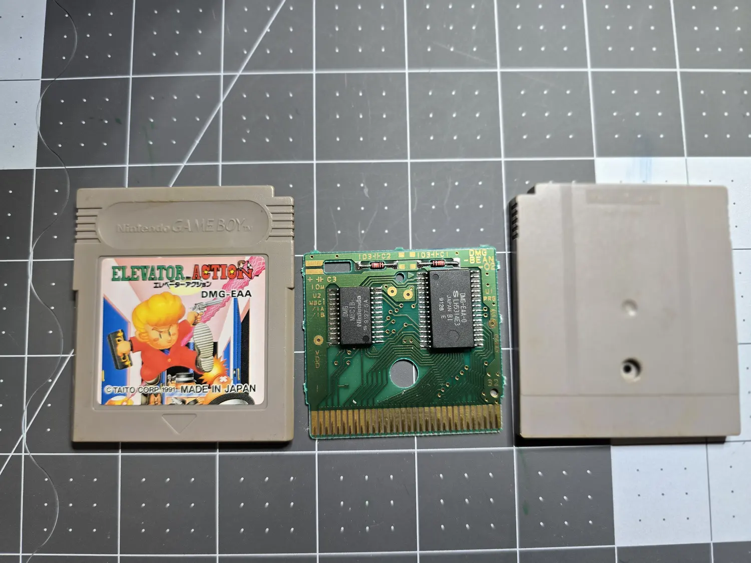 A Japanese Game Boy cartridge in three pieces on a cutting mat: front shell with an Elevator Action label reading DMG-EAA, copyright Taito Corp 1991, Made in Japan; the bare green PCB; and the grey back shell. The PCB is silkscreened DMG-BEAN-02 up the right edge and carries two black SOP chips, the left marked DMG MBC1B Nintendo S 9127 5A and the right marked DMG-EAA-0 S LH5314E3 Japan B1 9126 E, with the same U2 MBC1 slash 1A slash 1B silkscreen and a 32-pin gold edge connector.