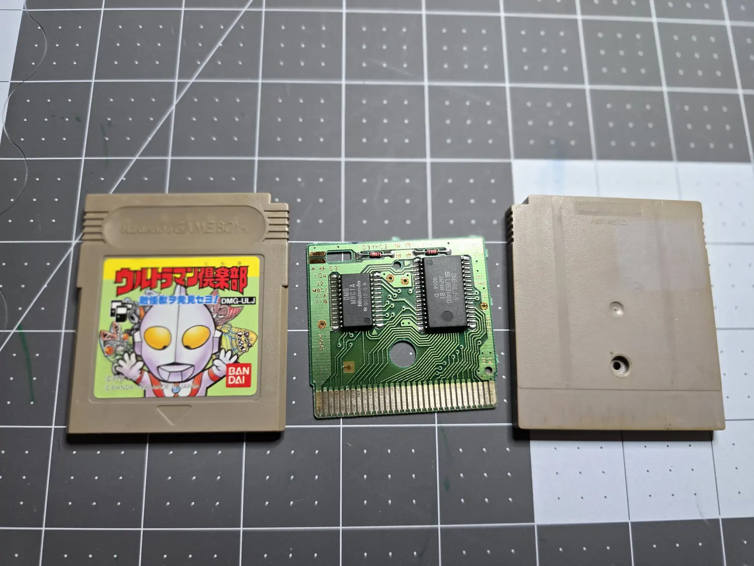 A Japanese Game Boy cartridge in three pieces on a cutting mat: front shell with a green Ultraman label reading Ultraman Club, DMG-ULJ, copyright Bandai 1990, Made in Japan; the bare green PCB in the middle; and the plain grey back shell marked Made in Japan, Pat. Pend. The PCB carries two black SOP chips, the left marked DMG MBC1A Nintendo S 9012 5A and the right marked DMG-ULJ-0 S LH5314H0 Japan B1 9014 D, with silkscreen reading U2 MBC1 slash 1A slash 1B, C3 10u, VDD and GND, above a 32-pin gold edge connector.