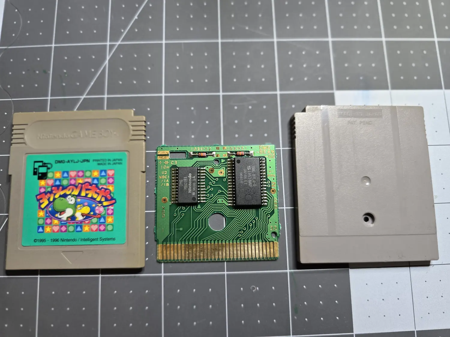A Japanese Game Boy cartridge in three pieces on a cutting mat: front shell with a green Yoshi label reading DMG-AYLJ-JPN, Printed in Japan, Made in Japan, copyright 1995 1996 Nintendo slash Intelligent Systems; the bare green PCB silkscreened DMG-BEAN-02; and the grey back shell. The PCB carries two black SOP chips, the left marked DMG MBC1B1 Nintendo S 9638 3A and the right marked DMG-AYLJ-0 S LH534MWF Japan E1 9640 D, above a 32-pin gold edge connector.
