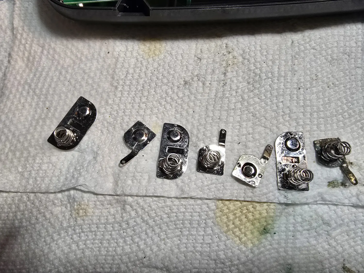 Seven Game Gear battery terminals laid out on a paper towel after cleaning, most now bright bare metal with the spring coils clean and shiny; the terminal at the far right is still visibly dark and corroded.