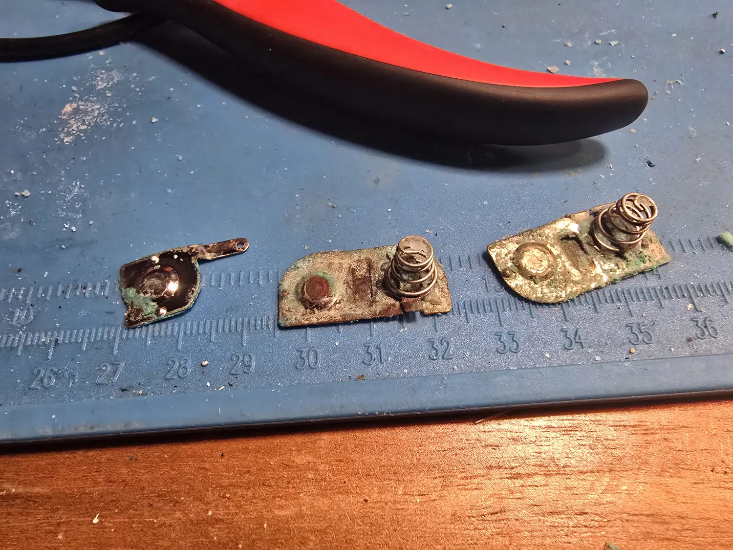 Three Game Gear battery terminals laid on a blue cutting mat with a millimetre scale, heavily corroded: bright green verdigris and white crystalline crust cover the springs and the flat contact plates, with the plating eaten away in patches.