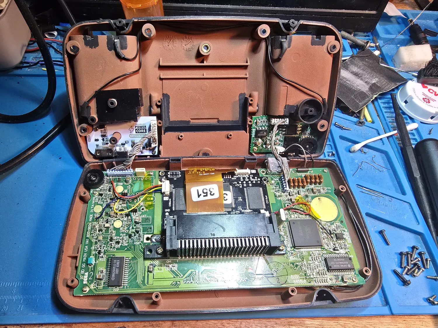 A Game Gear opened on a blue bench mat with the main board still in the brown rear shell. The board is silkscreened SEGA IC BD GG MAIN USA VA1 837-9024 Made in Taiwan along its lower edge. One large Sega QFP sits at the right-hand end beside a Hitachi 65256BLFP-10T SRAM dated Japan 9244, and a Samsung KM6264BLG-10 SRAM is at IC3 on the left. An aftermarket IPS driver board is fitted across the centre above the 45-pin cartridge connector, its orange ribbon carrying a white label marked 351. A small white board silkscreened POWER CORE, Version 1.0.1, Designed by RetroSix is mounted in the front shell at the upper left, with a second small green board at the upper right, both wired back to the main board with fine grey and coloured leads. Loose screws are laid out on the mat to the right.