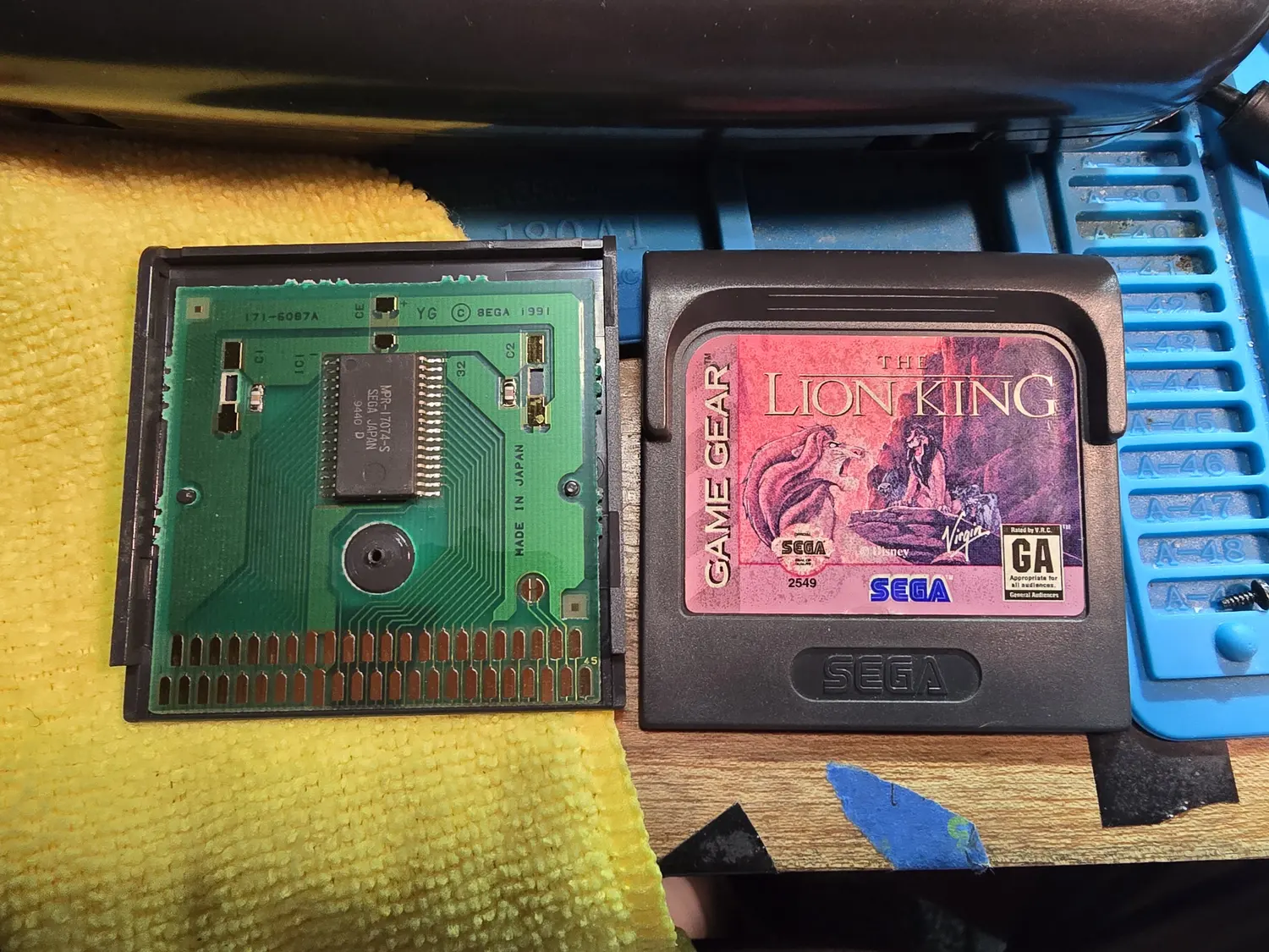 An opened Game Gear cartridge next to an assembled one. The exposed green PCB is silkscreened 171-6087A, YG, copyright SEGA 1991 and MADE IN JAPAN, carrying a single wide SOP mask ROM marked MPR-17074-S SEGA JAPAN 9440 D, with the 45-pin gold card edge along the bottom and the number 45 printed at its right end. The assembled cartridge beside it is a black Game Gear shell with a Lion King label.