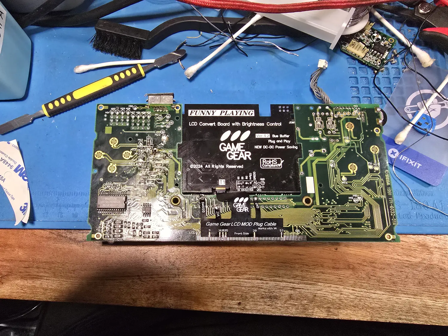 A Game Gear main board with a FunnyPlaying LCD replacement board fitted over its centre. The black add-on board is printed FUNNY PLAYING, LCD Convert Board with Brightness Control, Ver 4.2 Bus Buffer, Plug and Play, NEW DC-DC Power Saving, RoHS Compliant, copyright 2024. A flat plug cable below it is printed Game Gear LCD MOD Plug Cable, Works with V4, Front Side, with the numbers 9, 16, 18, 20, 39, 57 and 68 marked along its contacts, and labels GGSMS, VCC and GND at its ends.