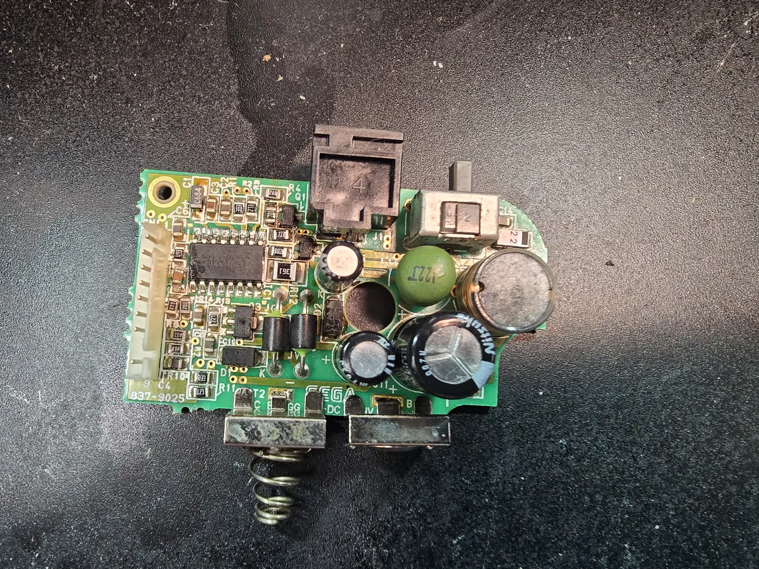 A Game Gear DC-DC power board silkscreened 837-9025 and SEGA, seen from the component side. A small surface-mount controller IC sits left of centre with surface-mount passives around it; several electrolytic cans of differing colours and a green disc capacitor sit to its right, along with a vacated circular footprint. The DC jack and the power slider are along the top edge, a white multi-way harness connector at the left, and two corroded battery terminals with springs project from the bottom edge.