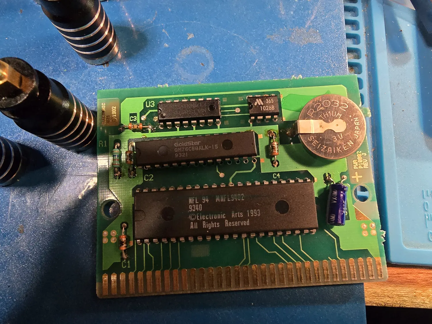 A Genesis cartridge PCB out of its shell, silkscreened PWA P1002 REV J and Electronic Arts 1992. It carries a large mask ROM marked NFL 94 MNFL9402 with a 1993 Electronic Arts copyright, a GoldStar GM76C88ALK-15 static RAM, a smaller 16-pin IC, and a Seizaiken CR2032 3 V lithium coin cell soldered flat to the board beside them, with the 64-pin gold card edge along the bottom.