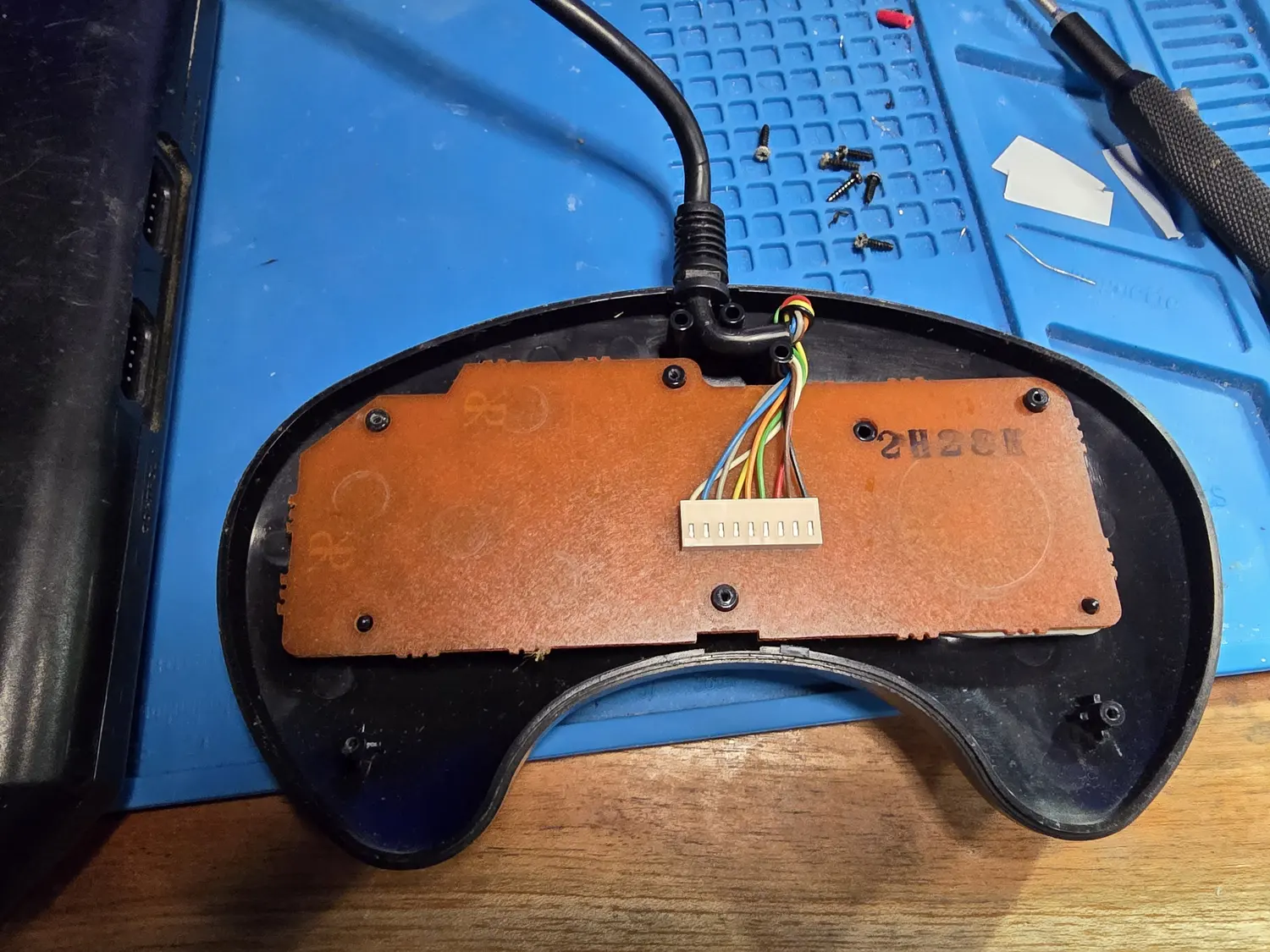 The back of an opened Genesis 3-button controller. A brown phenolic PCB fills the upper half of the black shell, held by several screws, with faint circular button outlines showing through. The cable enters through a moulded strain relief at the top and its nine coloured conductors run to a white multi-way crimped connector block plugged into the board.