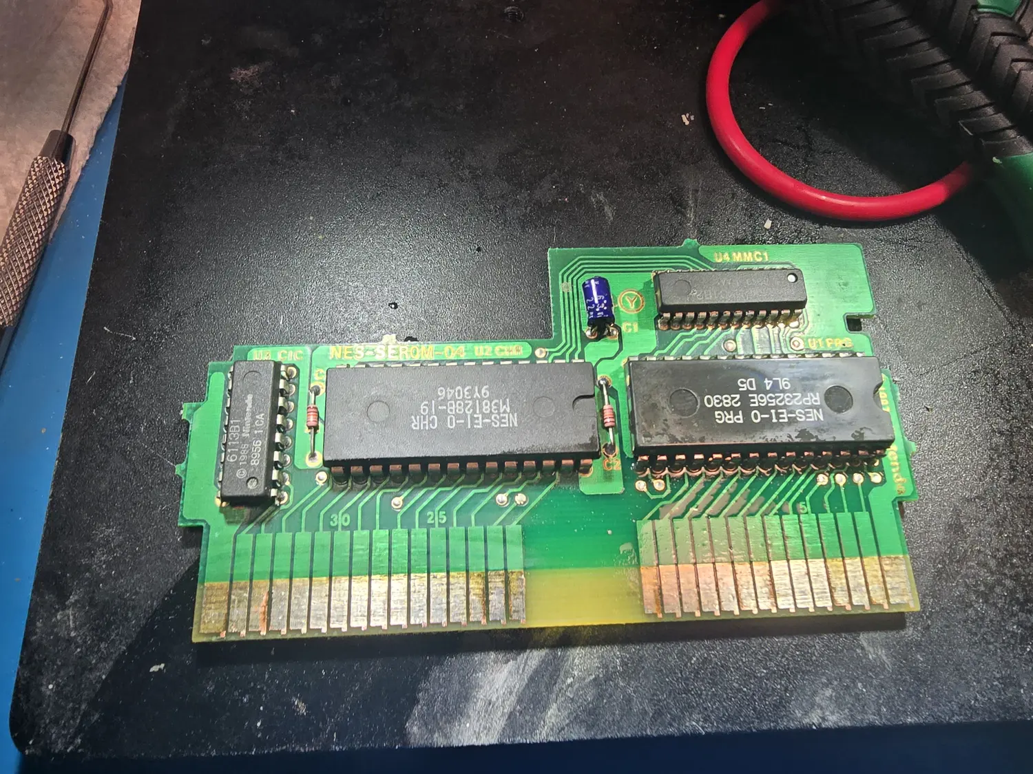 Component side of an NES cartridge board silkscreened NES-SEROM-04. Left to right it carries a 16-pin DIP marked 6113B1 with a 1985 Nintendo copyright at a position labelled U3 CIC, a 28-pin mask ROM marked NES-EI-0 CHR M38128B-19 at U2 CHR, a 28-pin mask ROM marked NES-EI-0 PRG RP23256E at U1 PRG, and a smaller DIP at U4 MMC1, with a blue electrolytic at C1. The 72-pin gold card edge runs along the bottom with silkscreened pin numbers 30, 25 and 5.