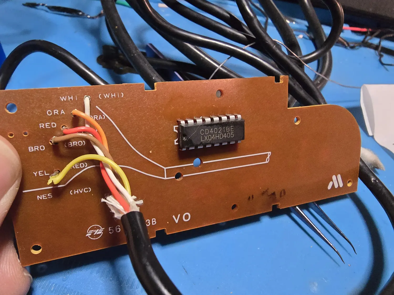 Solder side of an NES controller PCB on a brown phenolic substrate. A 16-pin DIP is marked CD4021BE over LX04HD405. At the left edge the cable conductors are soldered to pads labelled in two columns: WHI, ORA, RED, BRO, YEL and NES down one side, with WHI, ORA, BRO, RED and HVC bracketed alongside.