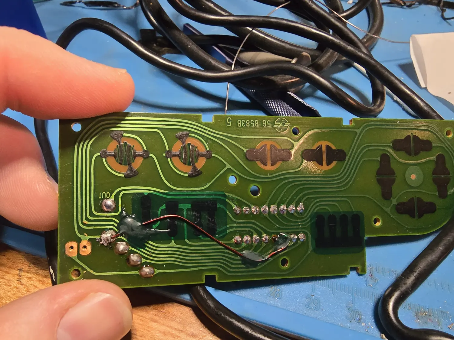 Contact side of an NES controller PCB, green, with the carbon contact pads for the D-pad, Select, Start and the A and B buttons. Silkscreen reads 56 85838 5. A fine red jumper wire runs diagonally across the middle of the board, its two ends covered by grey patches of solder mask, bridging from near a pad marked OUT across to the row of header solder points.