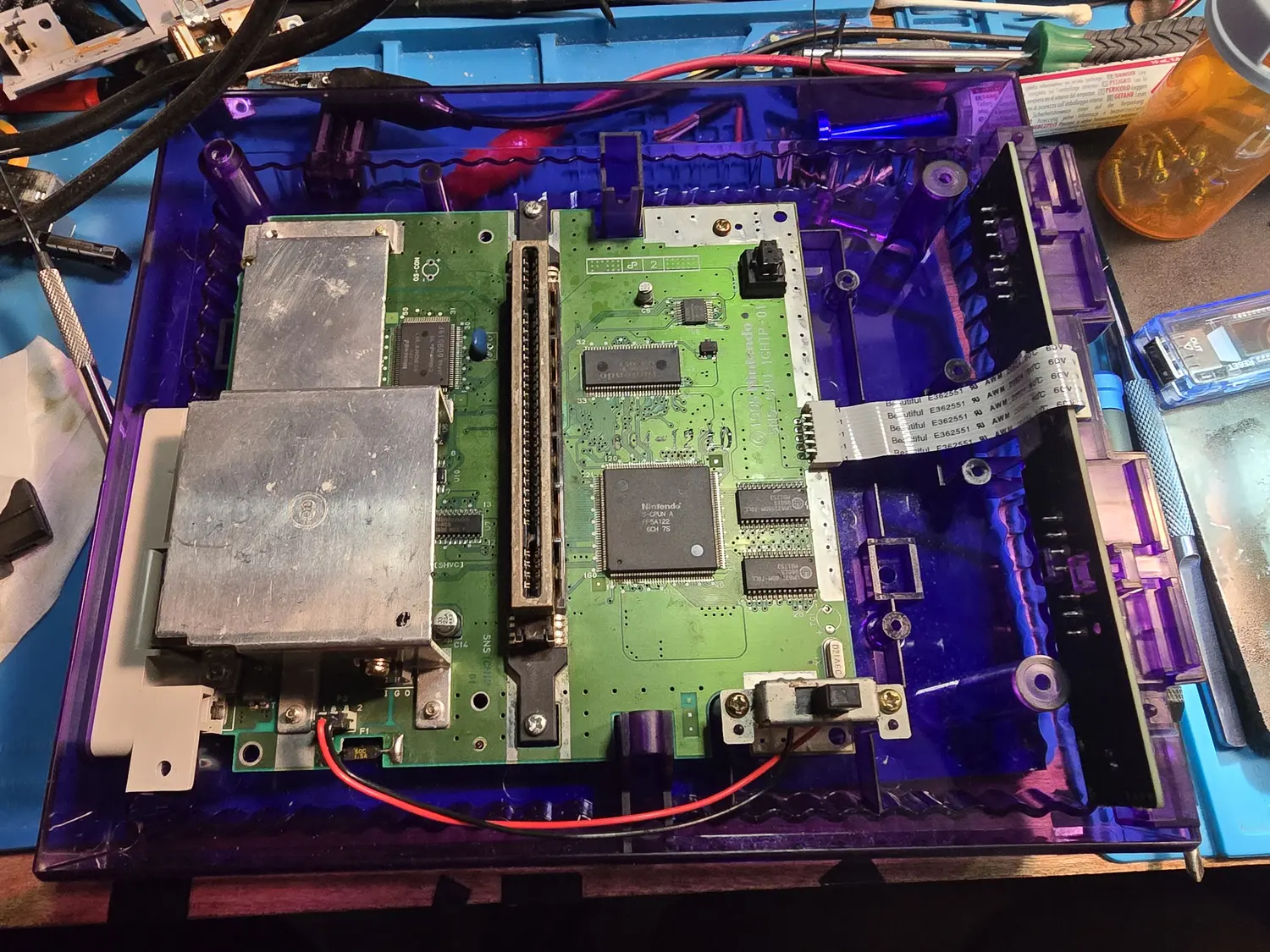A SNES mainboard fitted inside a translucent purple aftermarket shell, viewed from above. The board is silkscreened SNS-CPU-1CHIP-01 with a 1990 Nintendo copyright beside the 62-pin cartridge slot. A single large Nintendo QFP marked S-CPUN A FF5A122 sits to the right of the slot alongside two SMD RAM chips, with two metal RF shield cans covering the left half of the board and a white ribbon cable leaving the right-hand edge.