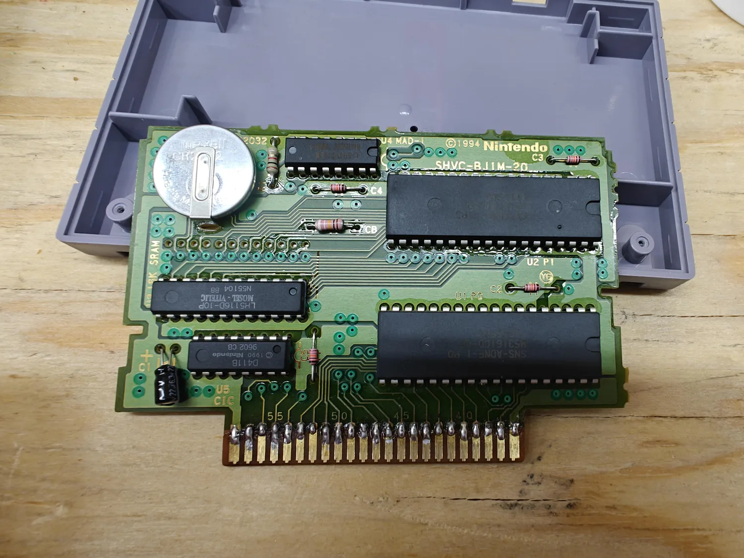 The same SNES cartridge PCB after repair, resting on the grey lower half of its cartridge shell on a wooden bench. The gold-finger edge strip is reattached to the board and every lane carries a bright solder joint bridging the board trace across the former break line onto its finger. The joints sit at the top of the finger area and the contact length below them is clean bare gold. Board silkscreen reads SHVC-BJ1M-20 and copyright 1994 Nintendo, with U4 MAD-1, U5 CIC, U2 P1 and U1 P0 designators and the finger-position numbers 55, 50, 45 and 40 visible above the joints.