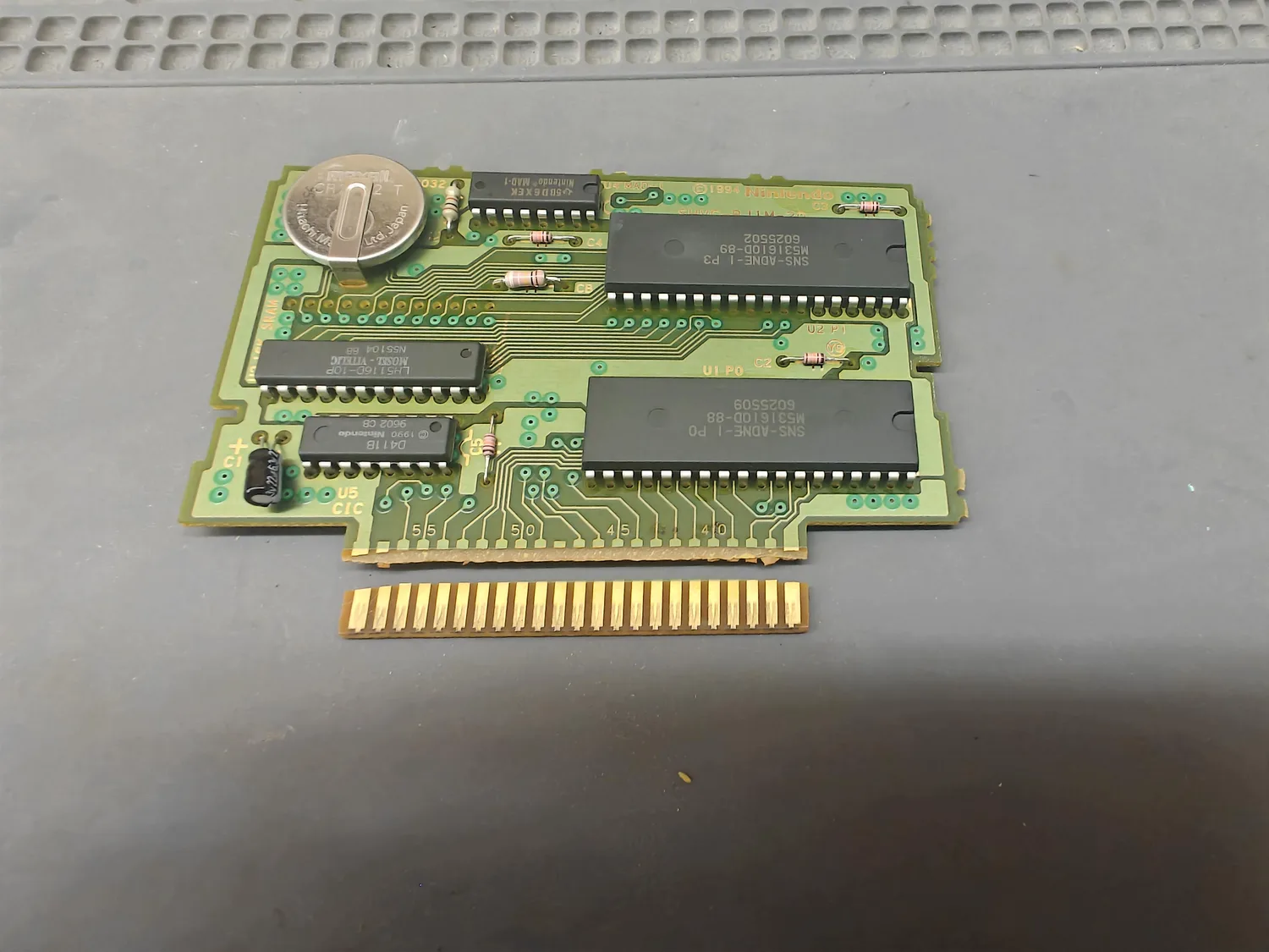 A green SNES cartridge PCB lying on a grey mat, photographed from the component side, with its entire gold-finger edge connector strip completely detached and lying loose below the board. The break runs the full width of the card edge, immediately below silkscreened finger-position numbers reading 55, 50, 45 and 40. The board carries a Maxell CR2032 coin cell in a holder at the upper left, a Sharp LH5116D-10P SRAM, a Nintendo MAD-1 at U4, a Nintendo D411B at U5 silkscreened CIC, and two black 40-pin mask ROMs marked SNS-ADNE-1 and MS31610D, with silkscreen reading SHVC-BJ1M-20 and copyright 1994 Nintendo.