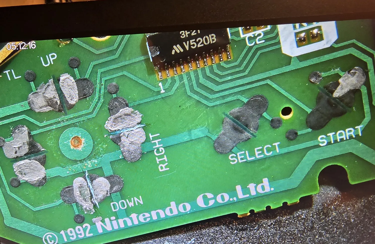 Microscope view of an SNES controller PCB. A small SOP chip at the top is marked 3F2? over V520B with a manufacturer logo, its pin 1 end marked with a 1 on the silkscreen. Silkscreen labels TL, UP, DOWN, RIGHT, SELECT and START sit beside carbon contact pads, several of which are covered with irregular grey and silver patches of conductive paint. The board is marked copyright 1992 Nintendo Co., Ltd.