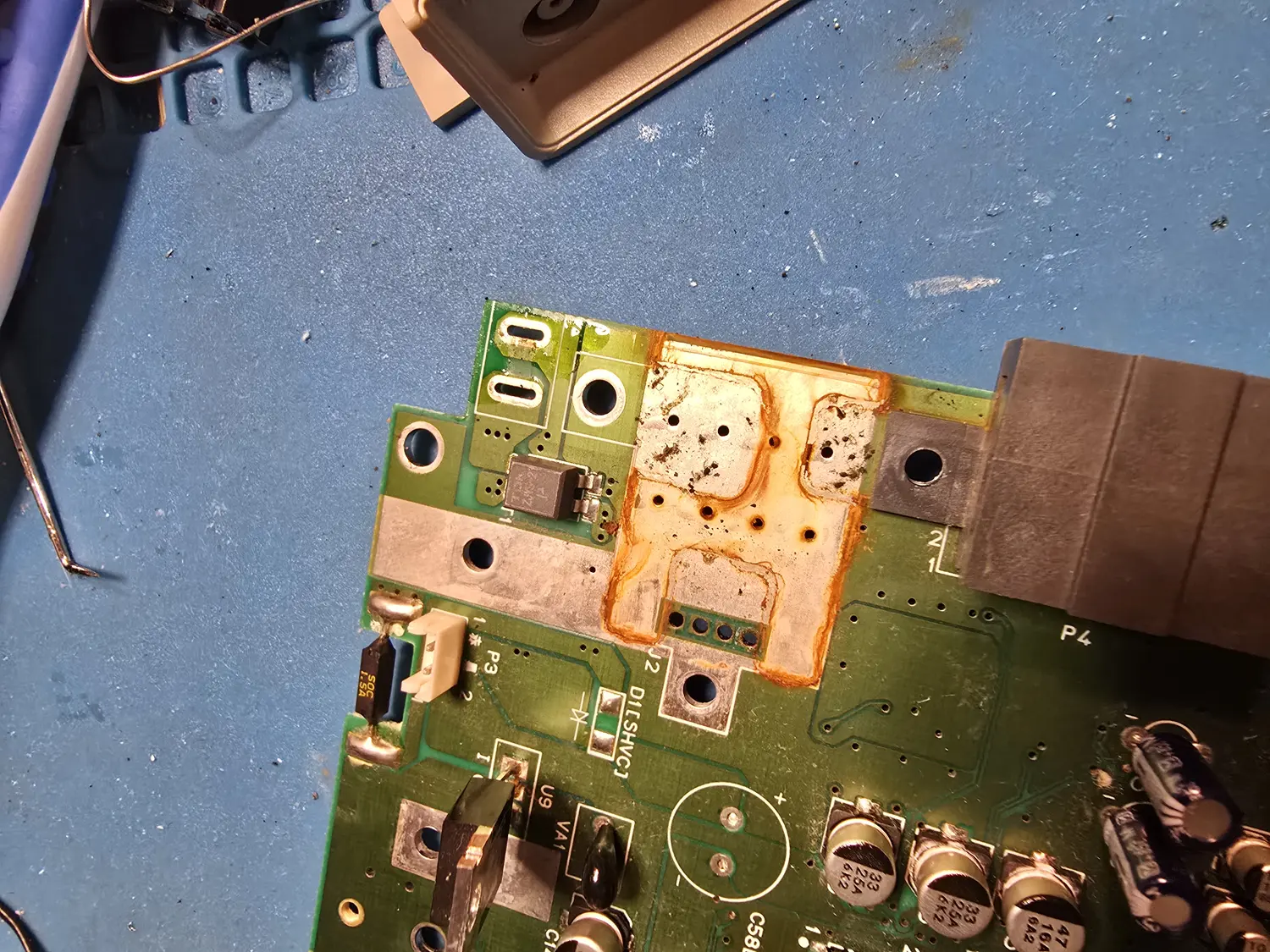 A SNES mainboard corner with the RF shielding removed and the DC barrel jack desoldered out, exposing a large rectangular area of orange-brown corrosion crust spreading across the solder mask, the module's mounting pads and the vacated jack footprint, which sits against large tinned ground areas. Nearby silkscreen reads P4, P3, J2, U9, VA1 and D1[SHVCJ], with a 1.5 A fuse and several electrolytic capacitors marked 33uF 25V and 47uF 16V visible at the lower edge.