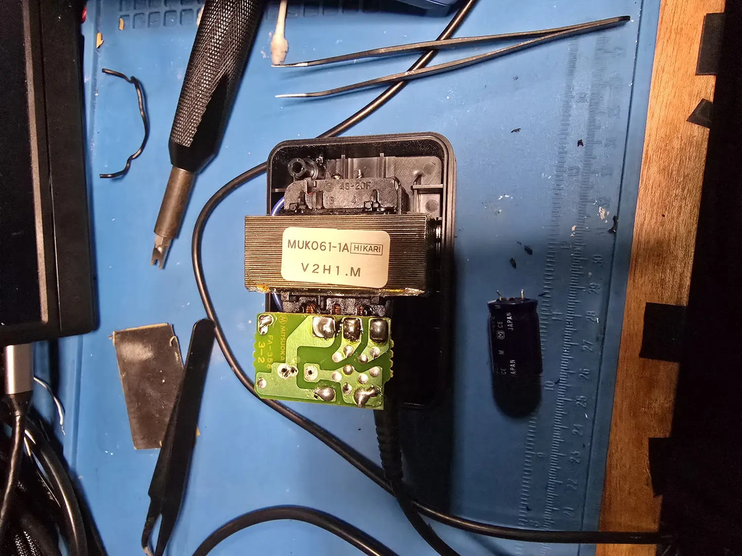 An opened OEM SNES power supply. Inside the black case sits a large laminated mains transformer with a label reading MUK061-1A HIKARI V2H1.M, above a small green rectifier board silkscreened MITSUOKA FA-358 3-2 carrying a handful of through-hole joints. A black axial electrolytic capacitor marked JAPAN lies on the mat beside the brick next to a steel rule.